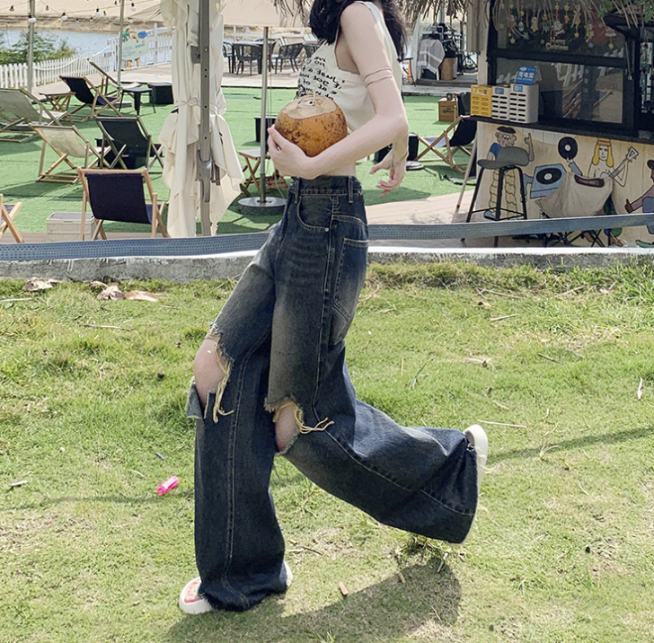 Side view of woman in casual wear holding coconut, wearing ripped jeans near beach seating area