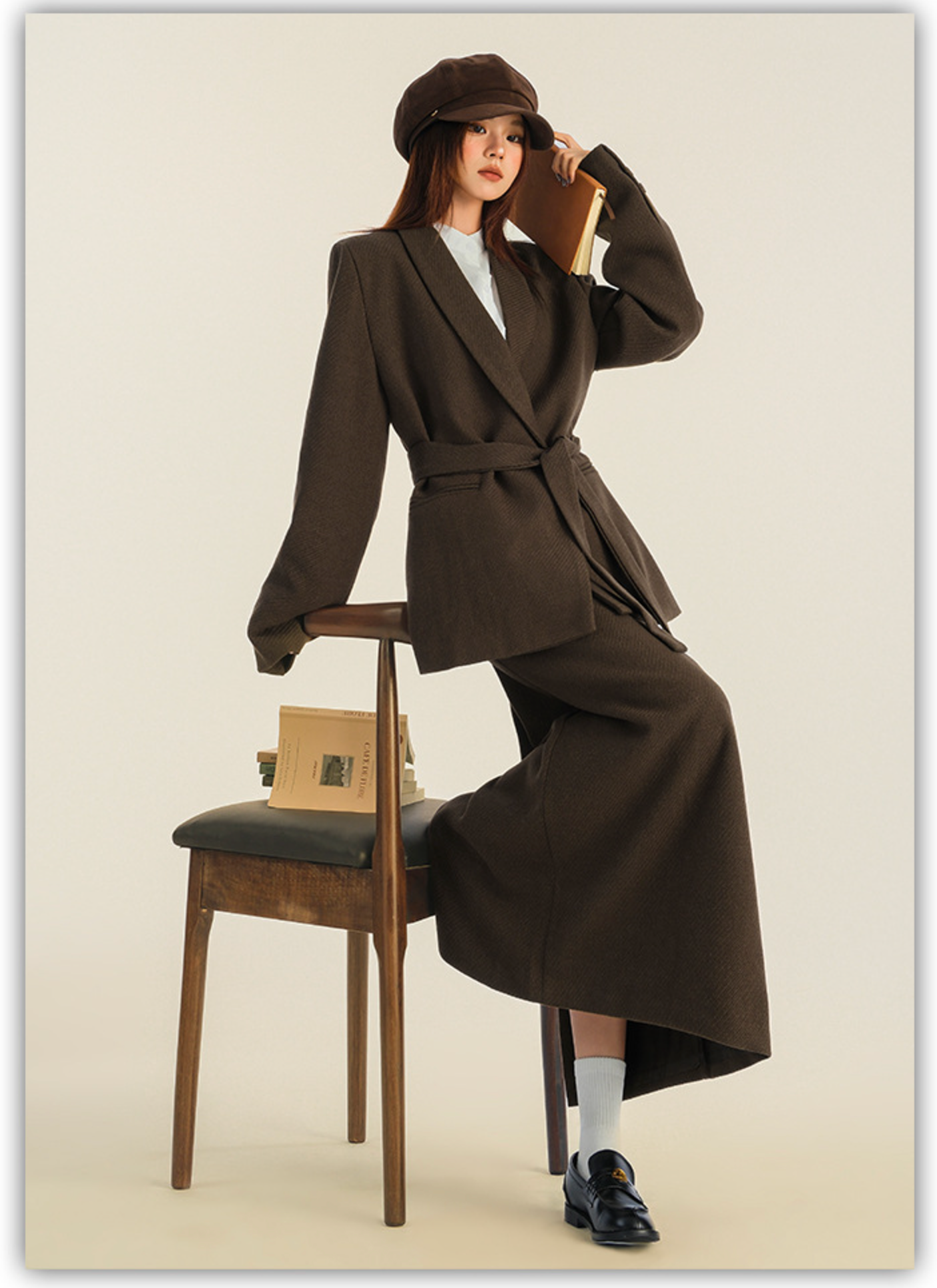 Model seated on wooden chair wearing brown wool suit set with newsboy cap and loafers