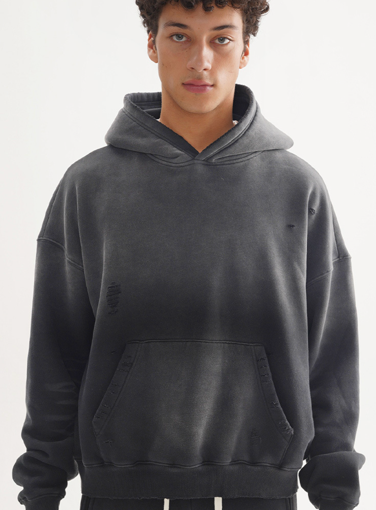 A model wearing charcoal grey oversized hoodie with distressed details and kangaroo pocket, posing against white background