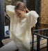 Woman in cream sweater adjusting ponytail against brick wall, wearing white pants
