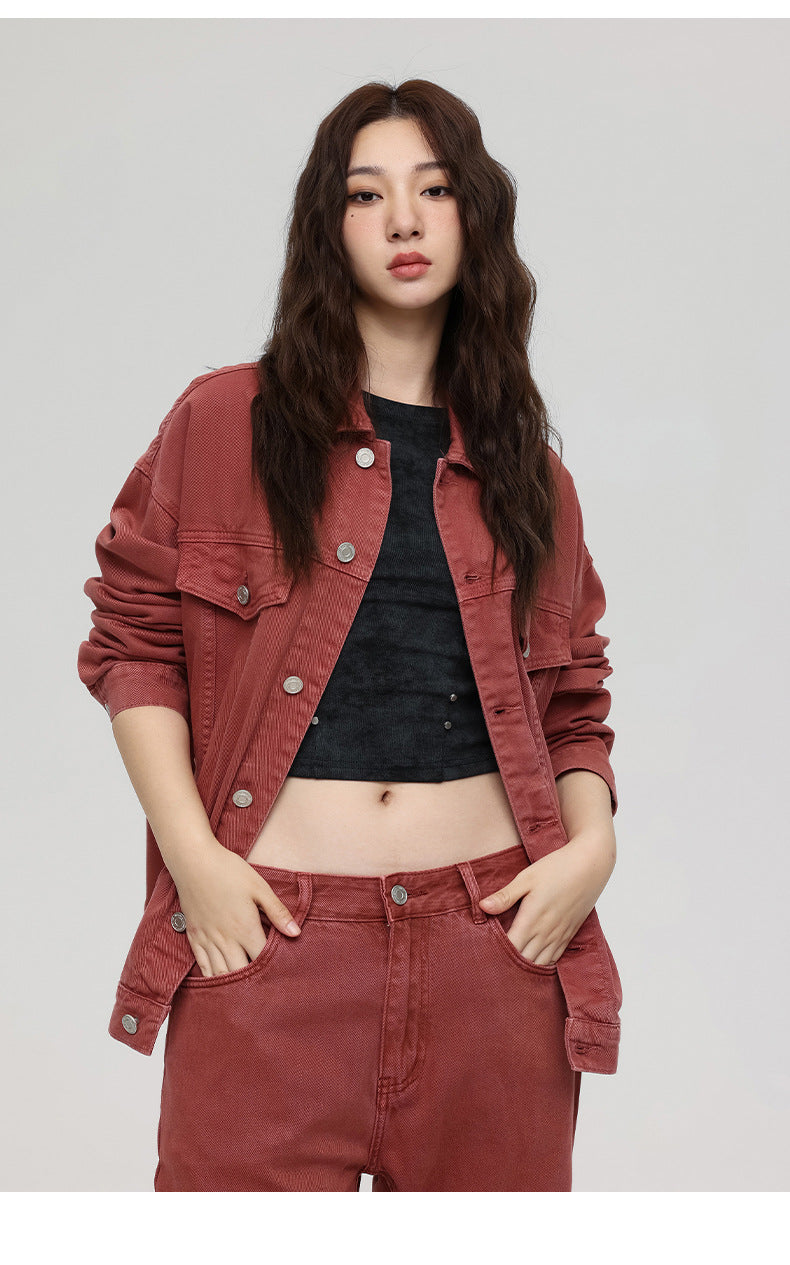 A woman with wavy hair poses in rust colored denim jacket over black cropped top showing midriff against white background