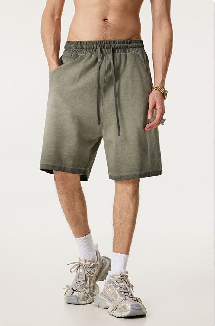 A man wearing olive green washed cotton shorts with drawstring waist, gold watch and white athletic socks against white background