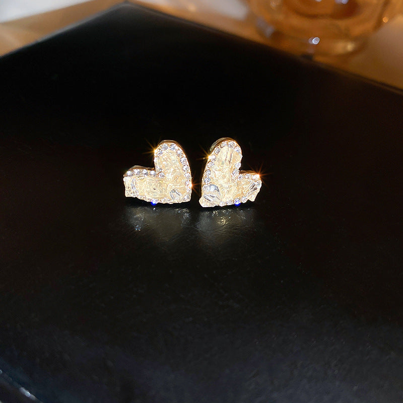  A pair of gold heart-shaped stud earrings with a textured surface and embedded zirconia, placed on a black background with soft lighting.