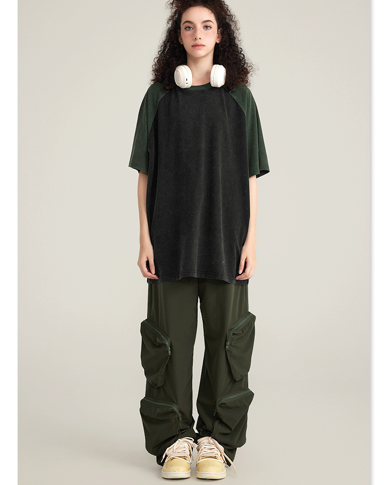 Model with curly dark hair wearing black and olive cargo pants with zippered pouches, oversized black/olive t-shirt, white headphones and cream sneakers on neutral background