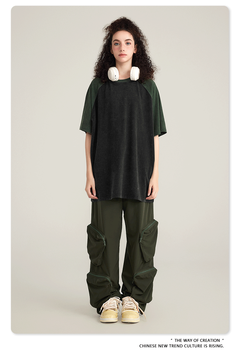 Model with curly dark hair wearing black and olive cargo pants with zippered pouches, oversized black/olive t-shirt, white headphones and cream sneakers on neutral background