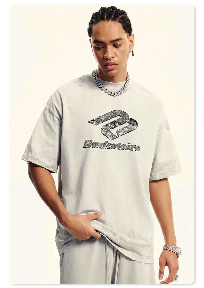 Close-up of model in cream Backstairs t-shirt with distressed logo print, silver chain necklace, hand in pocket against white backdrop