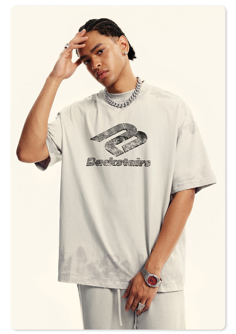 Model in cream Backstairs t-shirt with hand on forehead, silver chain necklace, ring and watch visible, distressed logo print against white background