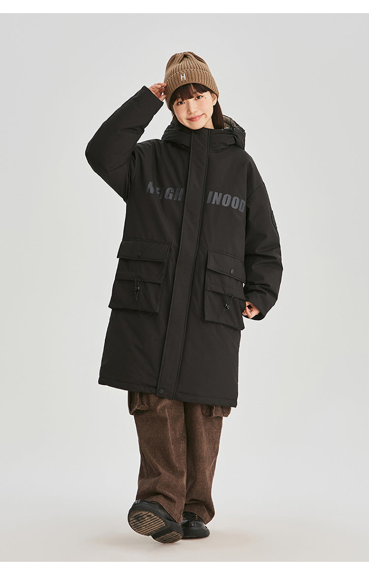 A woman wearing brown beanie adjusts her hat while modeling black NEIGHBORHOOD parka with brown pants against white background