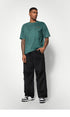 A male model wears a green oversized T-shirt, black wide-leg pants, and black-and-white sneakers, standing against a white background