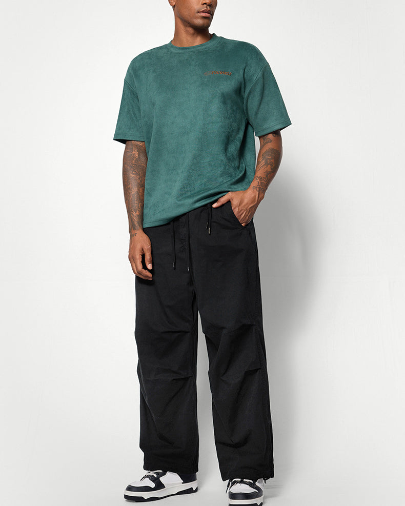 A male model wears a green oversized T-shirt, black wide-leg pants, and black-and-white sneakers, standing against a white background
