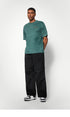 A male model poses in a green textured T-shirt and black wide-leg pants, looking slightly to the side