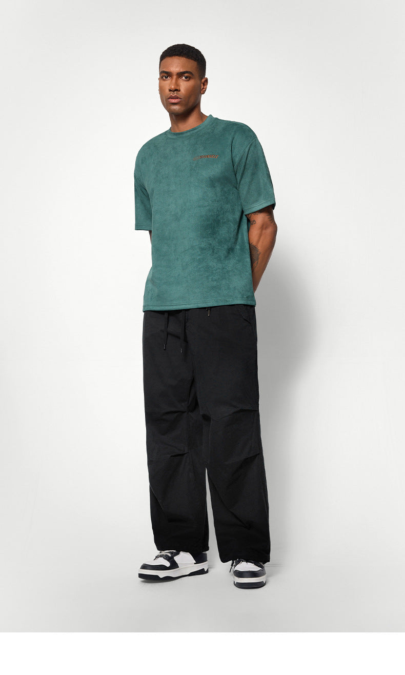 A male model poses in a green textured T-shirt and black wide-leg pants, looking slightly to the side