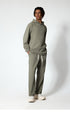 A full-body shot of a man in a sage green hoodie and sweatpants, wearing white sneakers, standing against a white wall