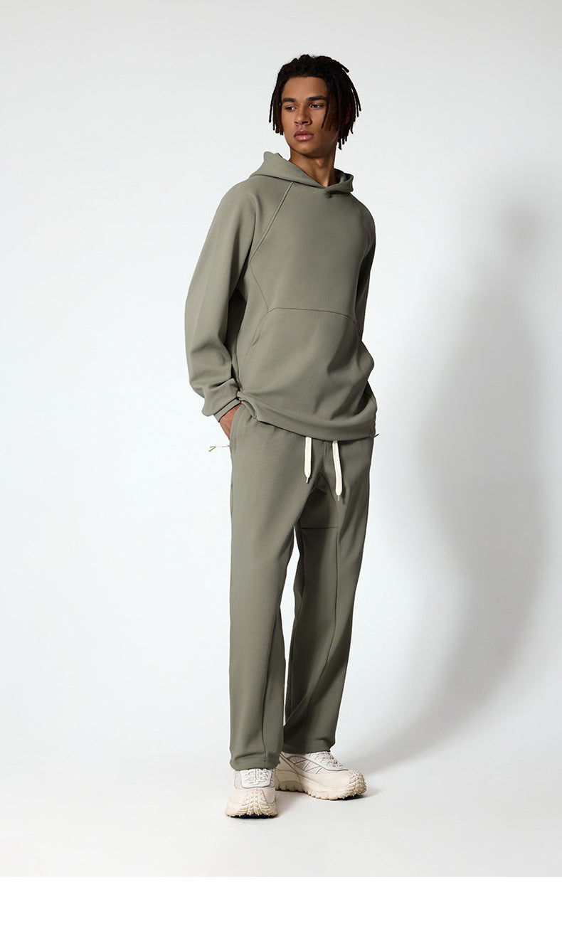 A full-body shot of a man in a sage green hoodie and sweatpants, wearing white sneakers, standing against a white wall