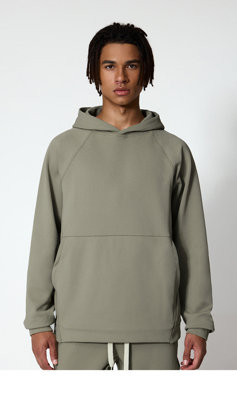 A man is wearing a sage green oversized hoodie with matching sweatpants, standing against a white wall
