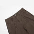 Close-up detail of brown ITKN pants showing front pocket construction and curved panel stitching against white surface