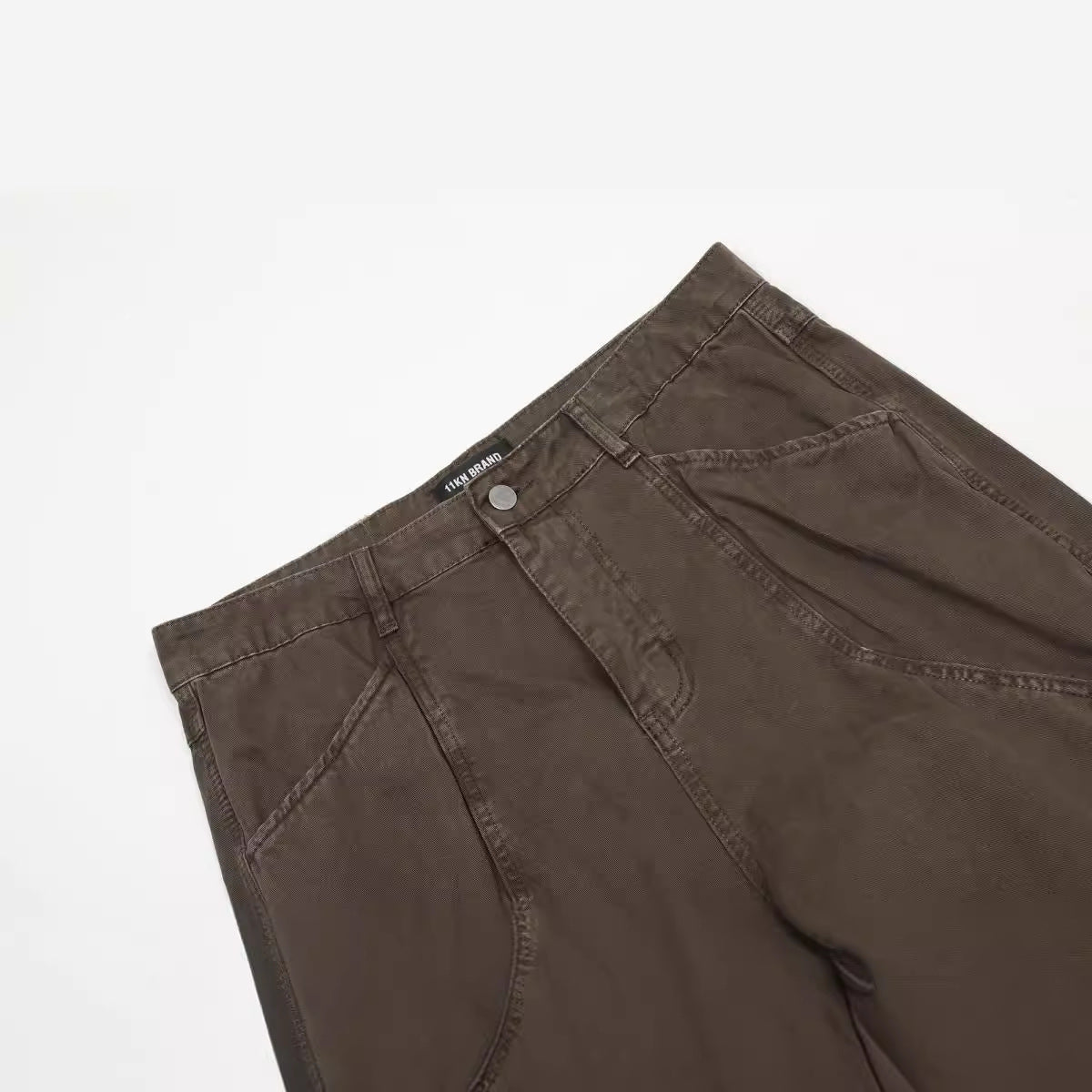 Close-up detail of brown ITKN pants showing front pocket construction and curved panel stitching against white surface