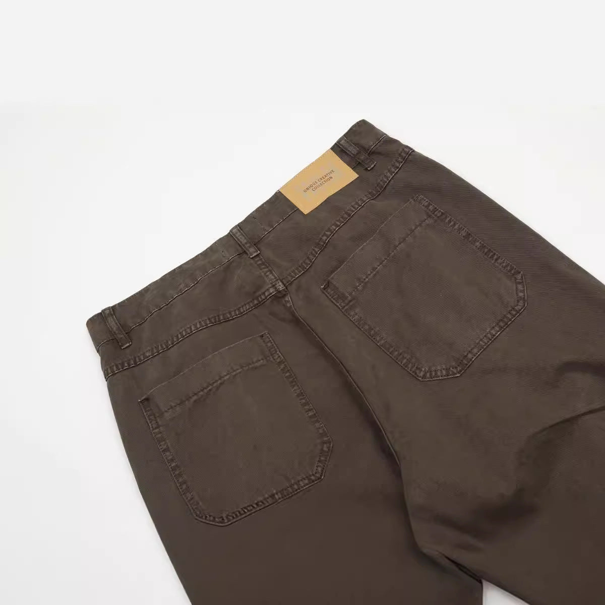 Back view detail of dark brown pants with patch pockets and brand label, photographed on white background