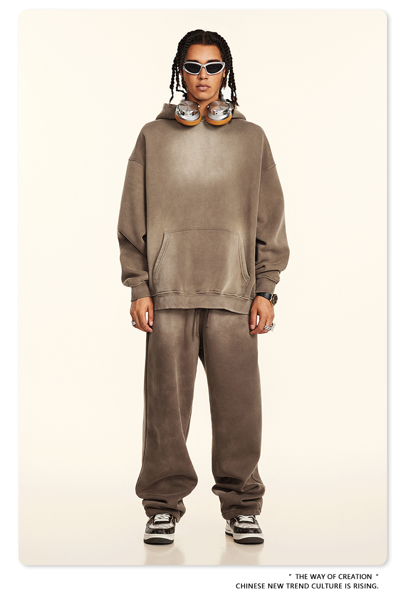 Model with twisted braids wearing taupe sweatsuit set with headphones around neck, standing against cream background