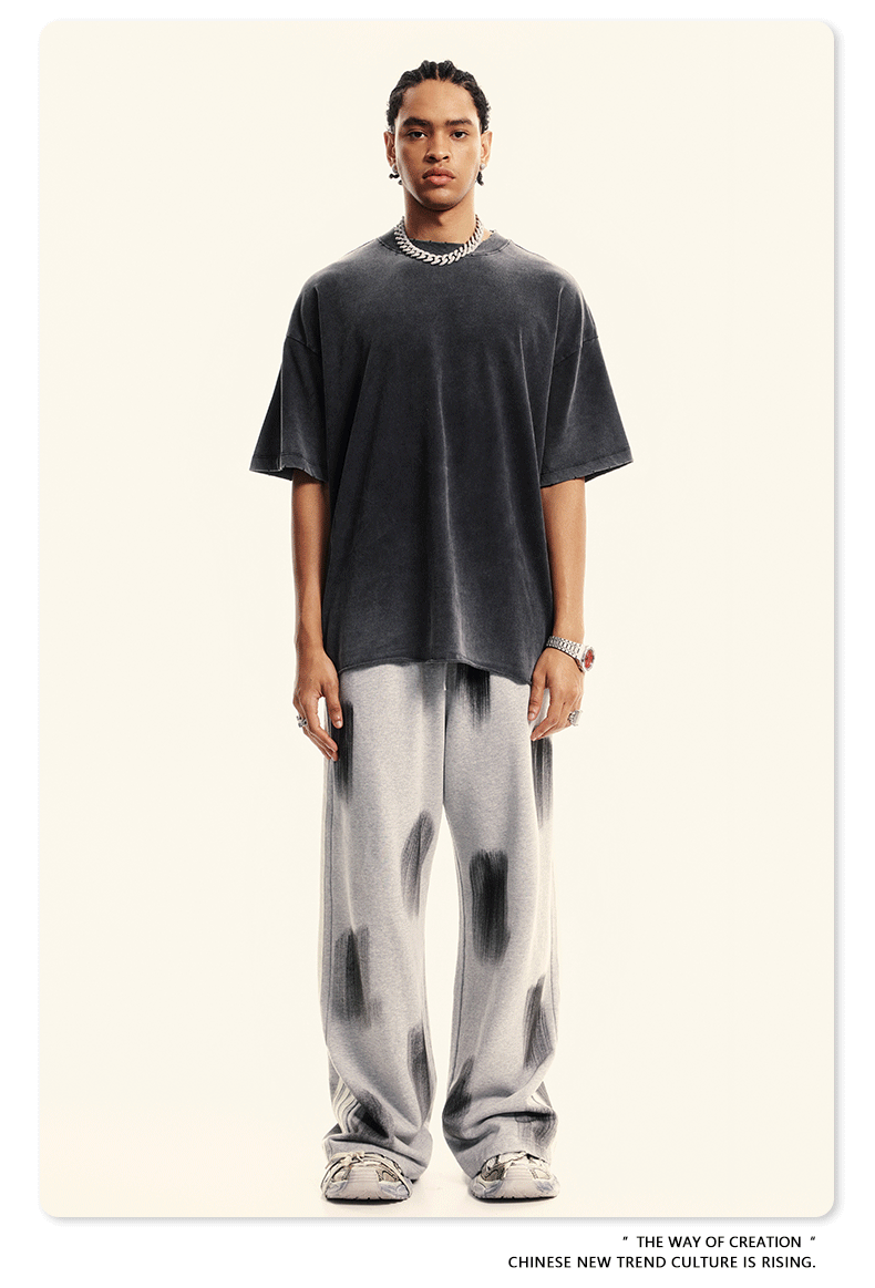 Model with braided hair wearing black oversized t-shirt, silver chain necklace, light gray pants with paint-like markings and white sneakers