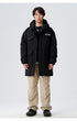A man wearing black Be_Born parka with silver chain necklace and beige cargo pants stands against white background