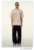 Model wearing beige/sand oversized t-shirt with white-framed sunglasses, silver chain necklace, black pants and gray sneakers against neutral background