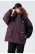 A woman with red curly hair adjusts her black Brooklyn cap while wearing plum/black All Drifting technical jacket against white backdrop