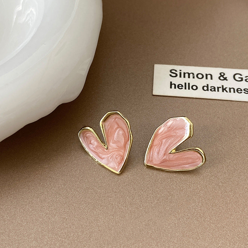 A pair of asymmetrical pink enamel heart earrings with a gold trim, set on a beige background beside a decorative card.