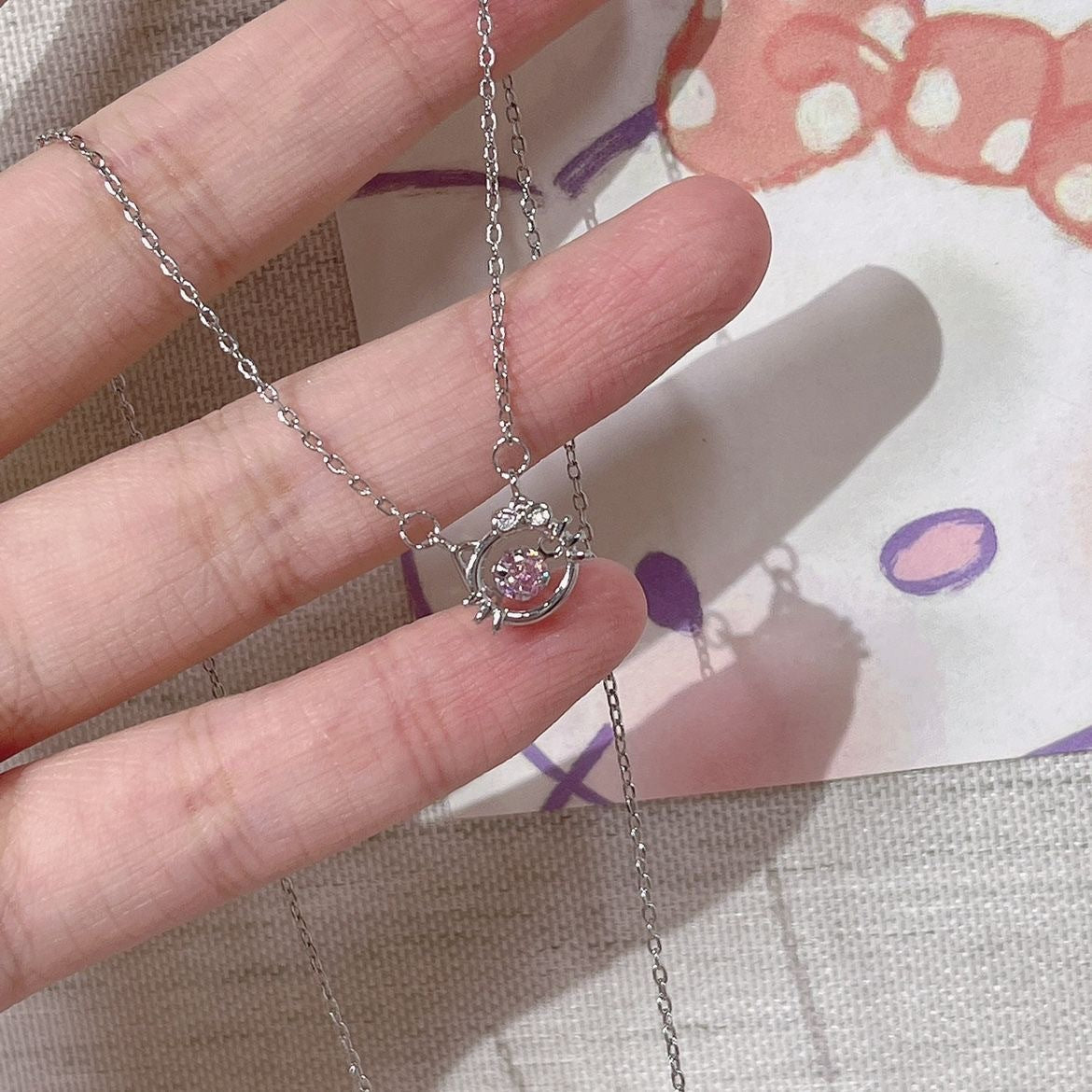 Close-up of silver cat pendant necklace with pink crystal center, held against patterned background