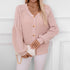 A model is wearing a pink button-up cardigan with puff sleeves holding a white clutch with gold chain, against white paneled wall background