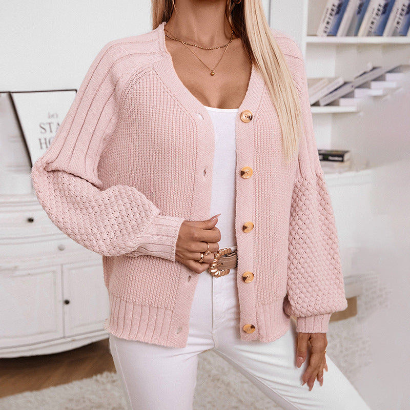A model is wearing a light pink ribbed cardigan with wooden buttons and textured puff sleeves, paired with white pants and gold accessories