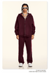 Model wearing burgundy oversized zip-up hoodie and matching sweatpants styled with white rectangular sunglasses and silver chain necklaces against cream backdrop