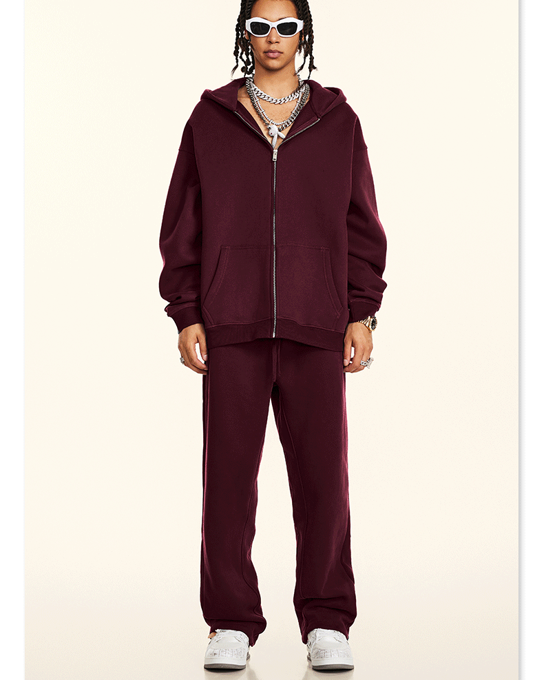 Model wearing burgundy oversized zip-up hoodie and matching sweatpants styled with white rectangular sunglasses and silver chain necklaces against cream backdrop
