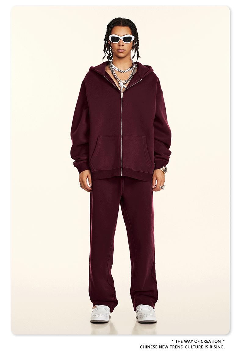 Model wearing burgundy oversized zip-up hoodie and matching sweatpants styled with white rectangular sunglasses and silver chain necklaces against cream backdrop