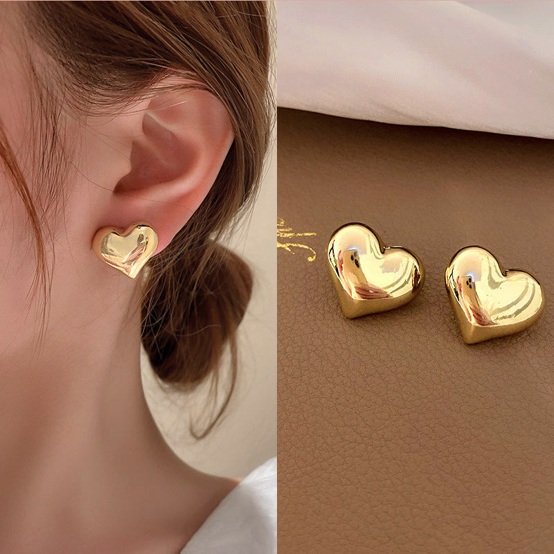 A pair of gold colored puffy heart stud earrings with a reflective, polished finish, shown both on a model and on a brown surface.