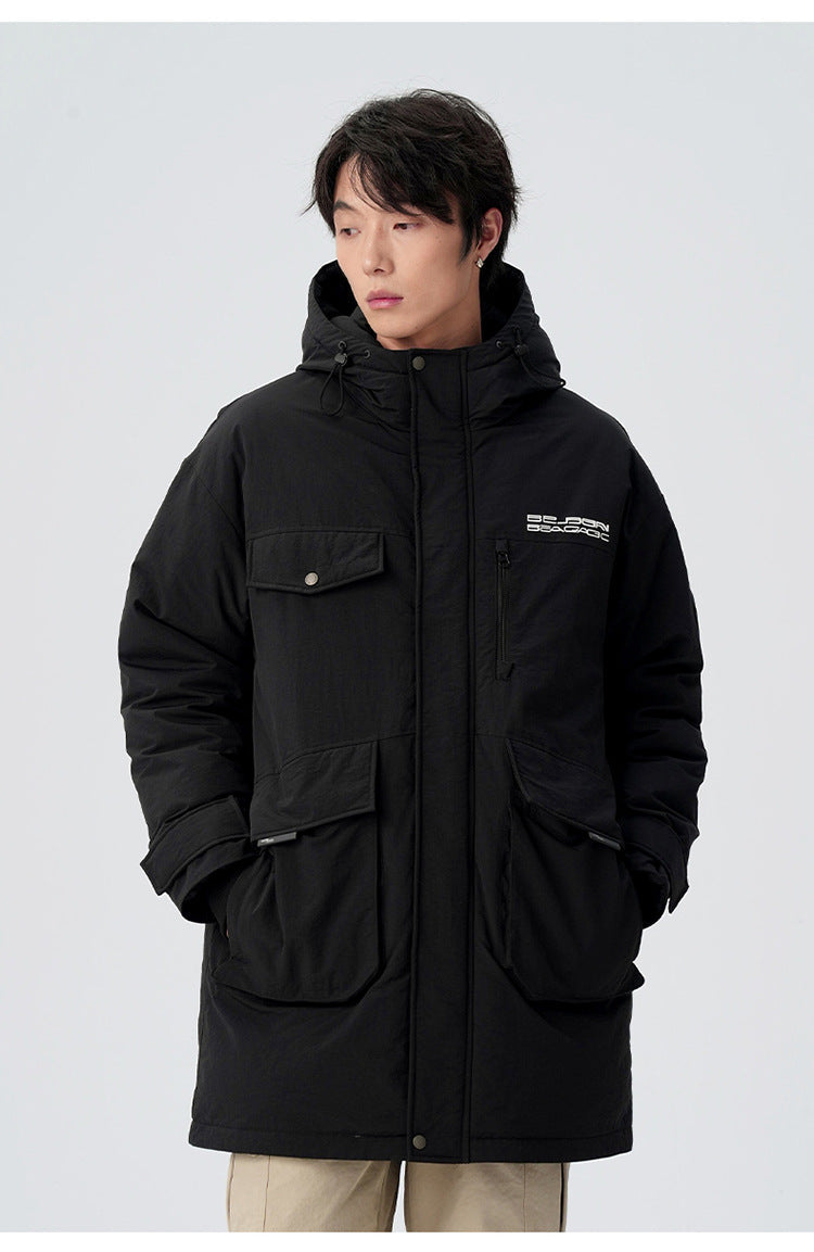 A man with hands in pockets wearing black Be_Born Beacaac parka with multiple utility pockets stands against white background