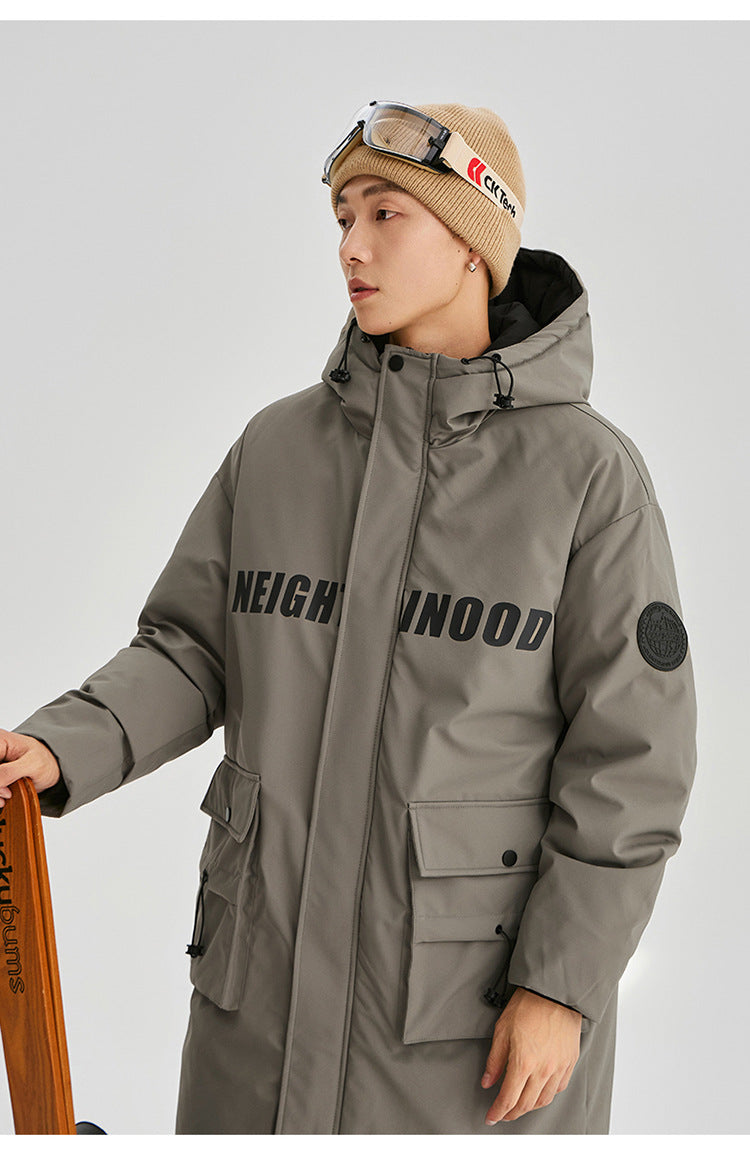 A man wearing beige beanie with ski goggles holds wooden snowboard while modeling olive NEIGHBORHOOD parka against white background