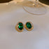 Gold stud earrings with oval green gemstones and black border displayed on white cushioned surface against neutral background.
