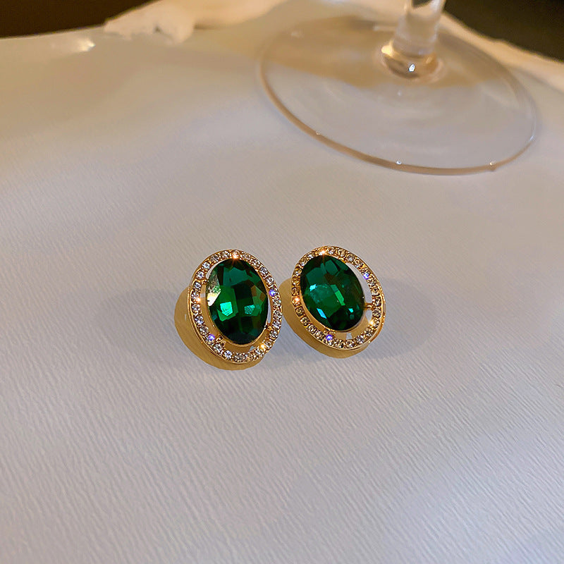 Gold stud earrings with oval green gemstones and black border displayed on white cushioned surface against neutral background.