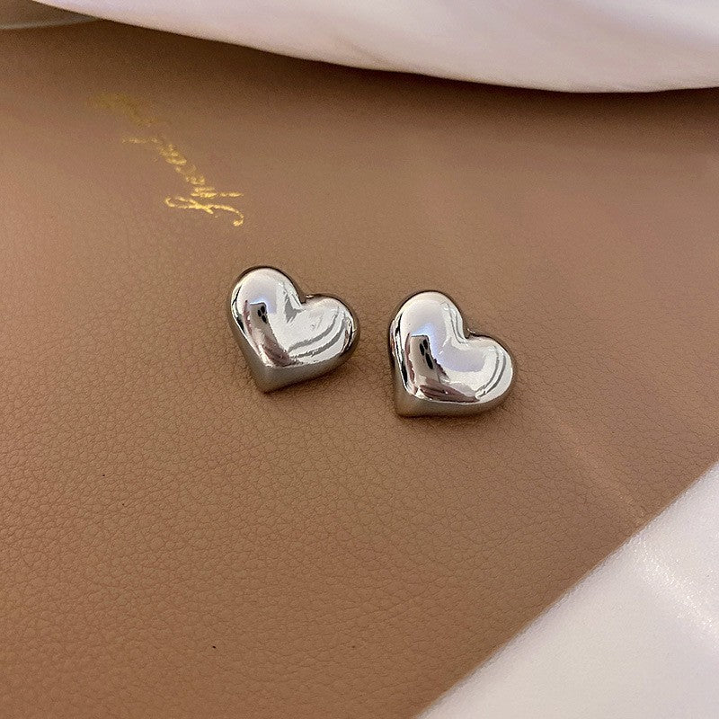 A pair of silver colored puffy heart stud earrings with a highly reflective, metallic finish, placed on a brown textured surface.