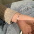 A woman is wearing a delicate silver heart charm bracelet with crystal accents on her wrist, paired with a cream sweater and blue jeans