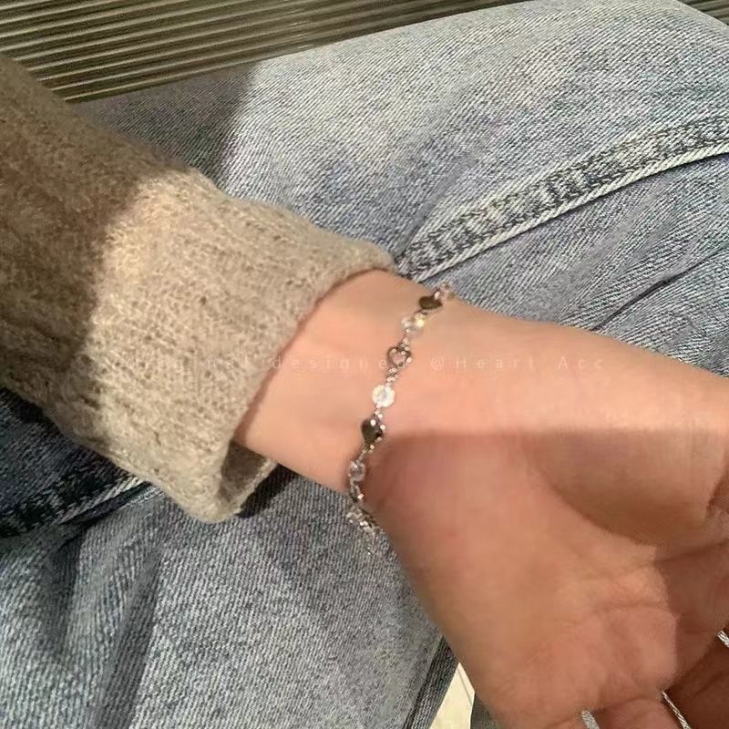 A woman is wearing a delicate silver heart charm bracelet with crystal accents on her wrist, paired with a cream sweater and blue jeans
