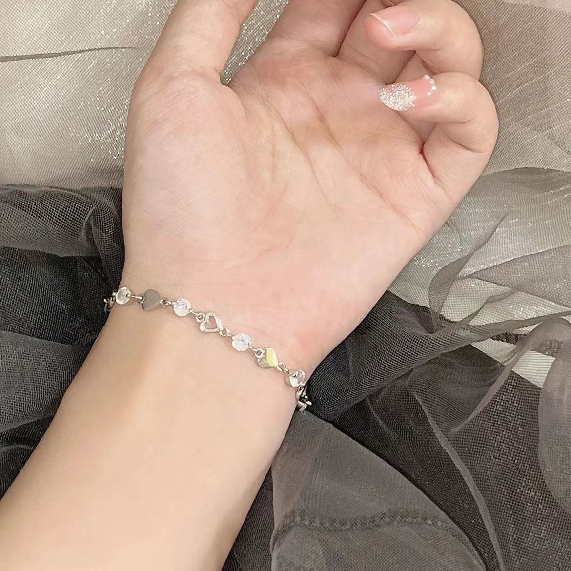 A woman is displaying a silver heart and crystal link bracelet on her wrist against sheer grey fabric background with soft lighting