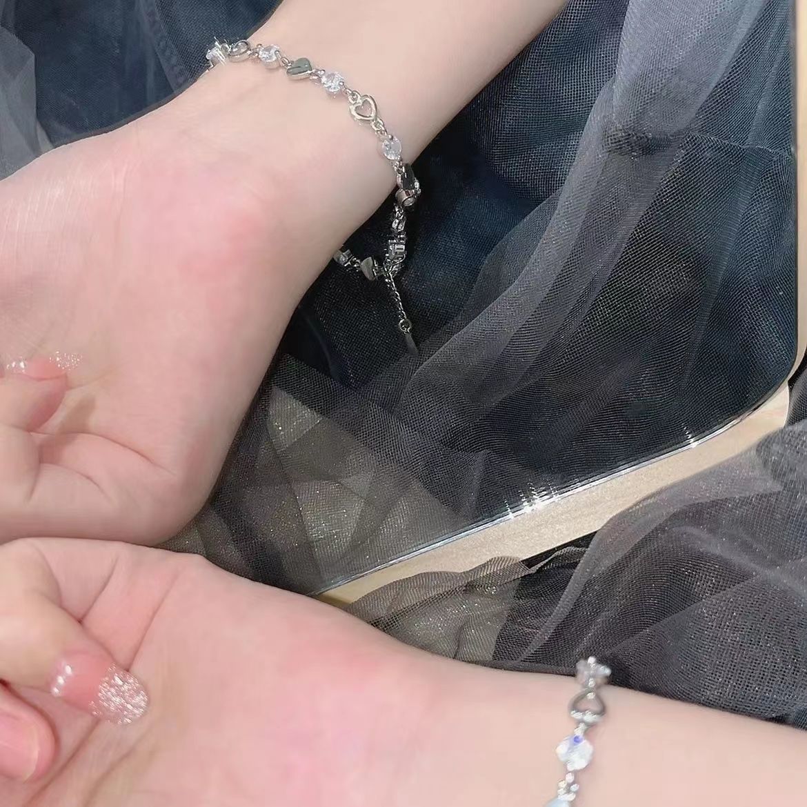 A woman is showcasing a dainty silver bracelet with alternating heart charms and crystals on her wrist against dark tulle fabric
