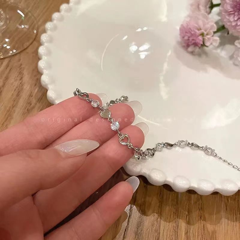 A woman is holding a silver heart and crystal chain bracelet with french manicure nails over a white scalloped plate with pink flowers