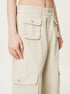 A close-up of beige cargo pants featuring buttoned flap pockets and a waistband with belt loops, worn by a model against a white background