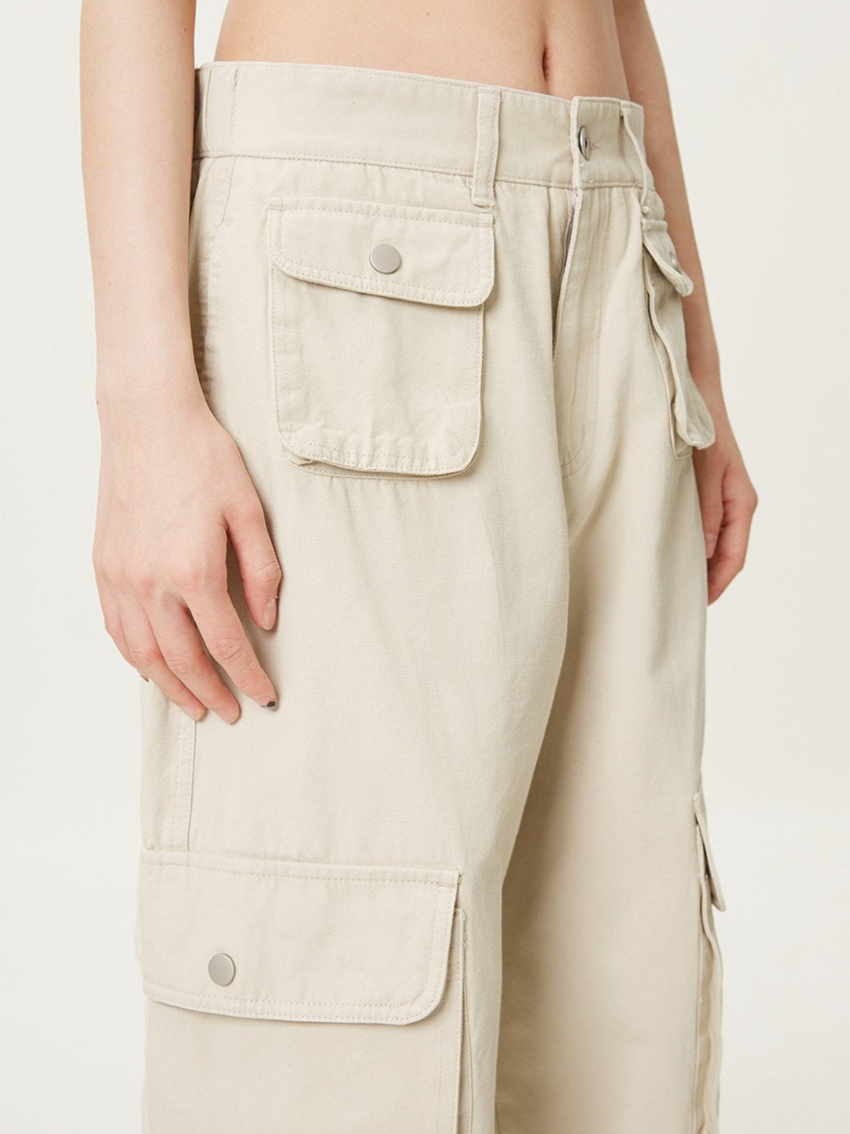A close-up of beige cargo pants featuring buttoned flap pockets and a waistband with belt loops, worn by a model against a white background