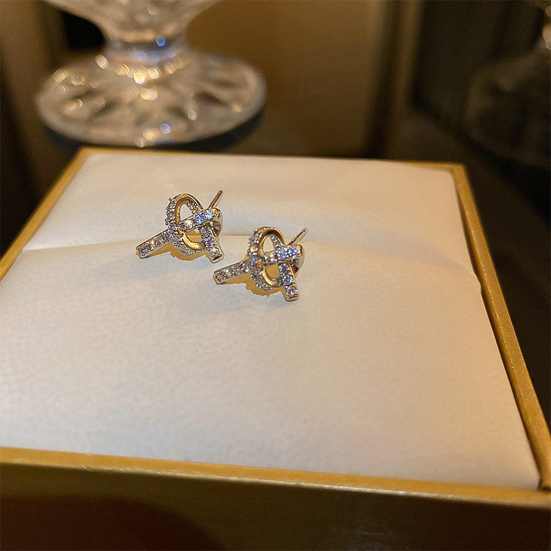 A pair of gold-tone bow-shaped stud earrings adorned with small cubic zirconia stones, displayed on a white cushion inside a gold-trimmed jewelry box.