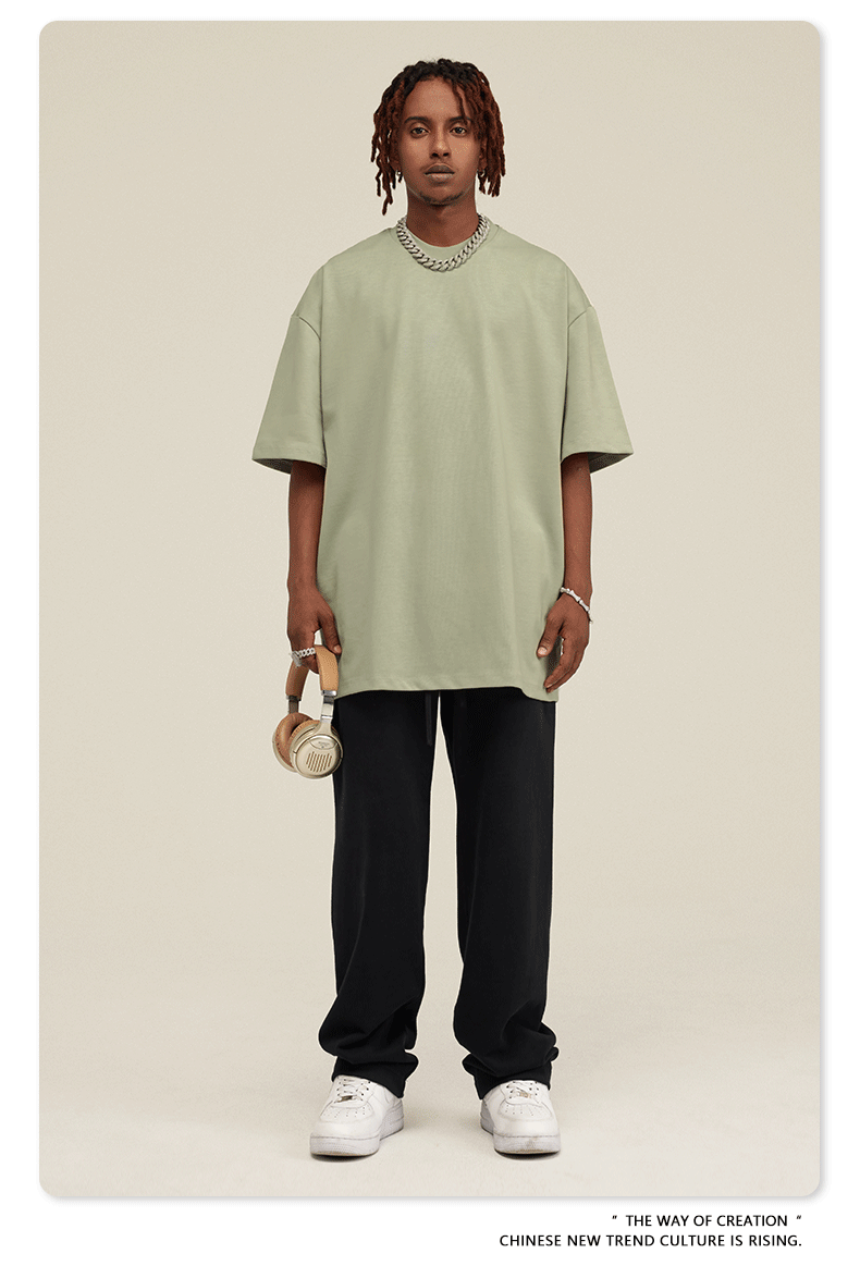 Man with short dreadlocks wearing light sage green oversized t-shirt, black sweatpants, silver chain necklace and white bracelet against neutral background