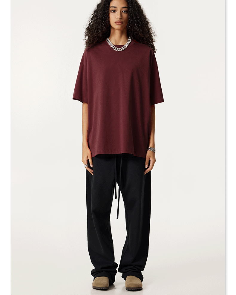 A model with long curly dark hair wearing a burgundy oversized t-shirt and black joggers, standing against a white background